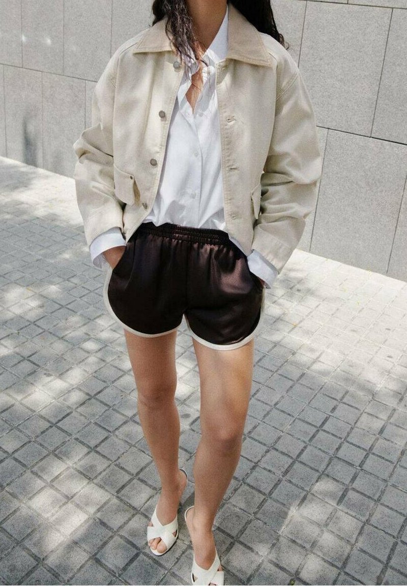 Personne debout sur une allée pavée portant une veste beige, une chemise blanche, un short de sport noir et des sandales blanches. Visage non visible.