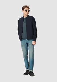 Giubbotto bomber navy sopra un maglione turchese, abbinato a jeans azzurri e scarpe nere; caratterizzato da un design minimalista e toni neutri.
