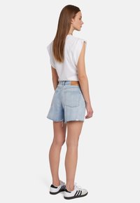 Top bianco senza maniche abbinato a pantaloni corti in denim blu chiaro. I pantaloni hanno un orlo sfilacciato e un'etichetta sulla vita. Le calzature sono sneakers nere e bianche.