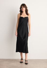 Robe midi en satin noir avec des bretelles fines, un décolleté festonné et une silhouette fluide. Associée à des escarpins noirs à bout pointu avec des accents dorés.