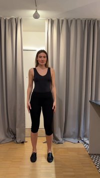 Kvinne står innendørs mellom beige gardiner, iført en svart ermeløs topp, svarte cropped bukser og svarte loafers på tregulv.