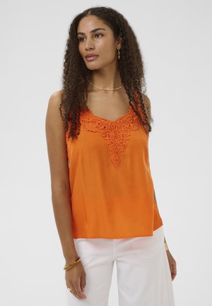 Kitsaste tumedate lokkidega naine, kellel on seljas oranž pitsdetailidega varrukateta top ja valged püksid, seisab ühevärvilise tausta ees.