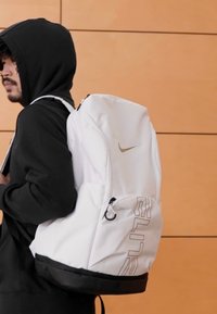 Bijeli Nike ruksak glatke teksture, zlatnog logotipa, bočni džep na patentni zatvarač i crno dno, nošen preko ramena od strane osobe.