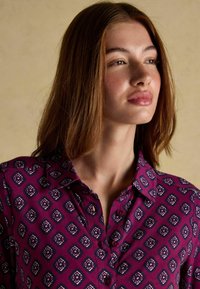 Fialová košeľa s gombíkmi s geometrickým vzorom a bielymi a modrými akcentmi. Dlhé rukávy, golier a mäkká textúra látky.