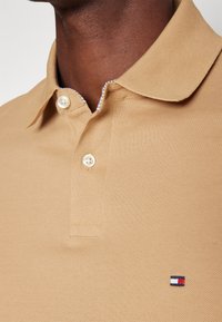 Beige pólóing textúrált anyagból, klasszikus gallérral, két gombbal és egy kis piros, fehér és kék logóval a mellkasán.