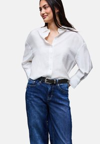 Bílá oversized košile na knoflíky s volným střihem, spárovaná s tmavě modrými širokými džínami a černým páskem, s hladkým povrchem.