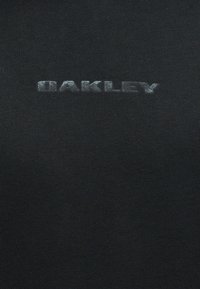 Αχνό λευκό λογότυπο "OAKLEY" τυπωμένο σε μαύρο υφασμάτινο φόντο με υφή.