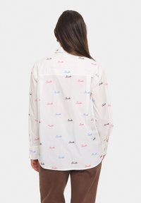 Camicia bianca a maniche lunghe con testo "Smile" sparse in rosa, blu e nero. Tessuto morbido con vestibilità rilassata e polsini abbottonati.