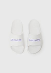 Sandales en caoutchouc blanches à semelle ouverte et talon ouvert, avec "LACOSTE" imprimé en violet sur le dessus. Semelle texturée pour une meilleure adhérence.