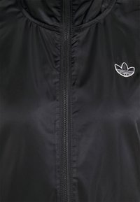 Svart huva jacka med slätt, glänsande tyg. Har en helängdsdragkedja och broderad logotyp på vänster bröst.