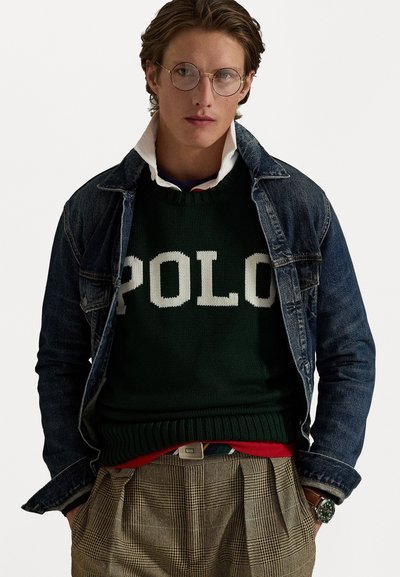 Groene gebreide trui met "POLO" tekst, gedragen over een witte bloes met kraag, bovenop een denim jack; gecombineerd met gestreepte bruine broeken.
