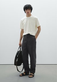 Camicia bianca a maniche corte con un leggero pannello sul petto, pantaloni grigi scuri a vestibilità rilassata, sandali neri e una borsa marrone con hardware argentato.