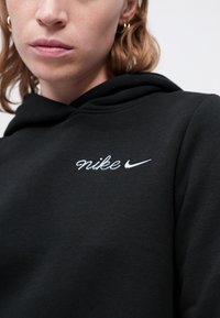Melnā Nike kapuce ar mīkstu tekstūru, kurai ir balts izšūts logos uz krūšu daļas. Dizainā ir iekļauta šnore vienādas formēšanas kapucē un brīvs griezums.