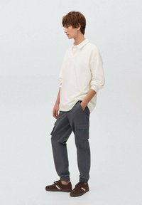 Valkoinen pitkähihaisen polo-paita, harmaat cargo-housut sivutaskuilla ja ruskeat tennarit valkoisilla yksityiskohdilla, seisoo tasaisella taustalla.