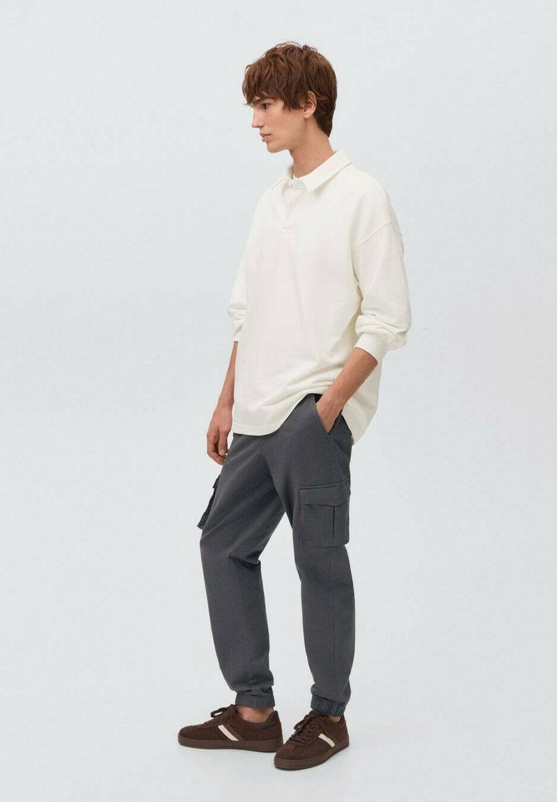 Cămașă polo albă cu mânecă lungă, pantaloni cargo gri cu buzunare laterale și adidași maro cu accente albe, stând în fața unui fundal simplu.