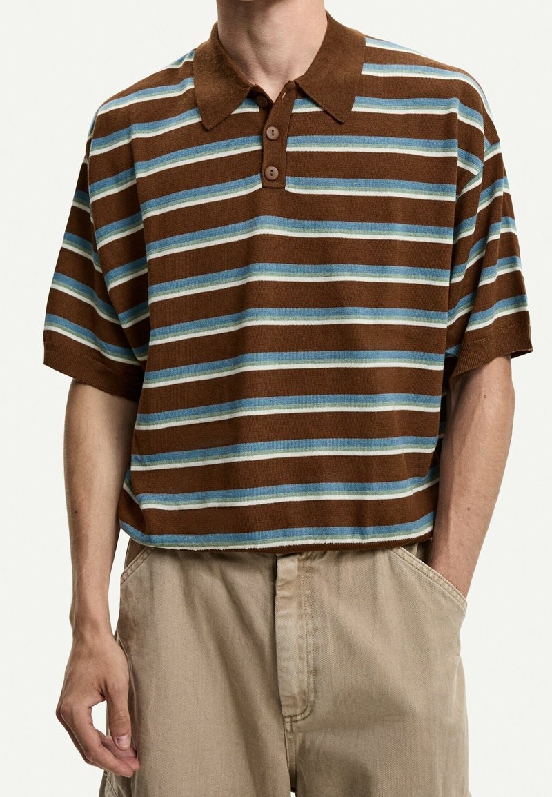 Homme portant un polo à manches courtes à rayures horizontales marron, bleu et blanc, pantalon beige, mains dans les poches, debout devant un fond blanc.