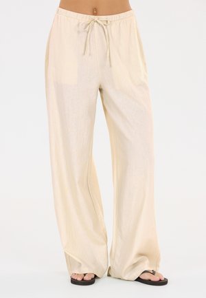 Løse beige bukser med elastisk talje og snøre, båret med sorte flip-flops, vist fra talje til fødder på en lys baggrund.