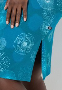 Tela azul verdoso con patrones florales y de puntos blancos, con una abertura lateral y una etiqueta negra. Textura suave, diseño informal.