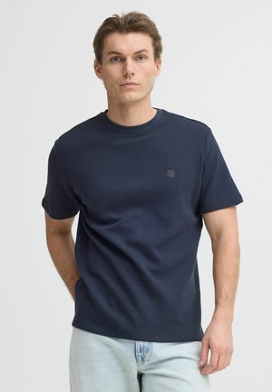 Junger Mann mit heller Haut und hellbraunen Haaren, trägt ein dunkelblaues, strukturiertes Kurzarmhemd und helle blaue Jeans, steht vor weißem Hintergrund.