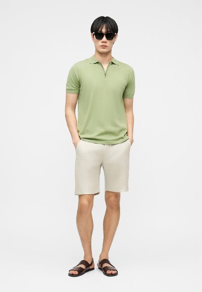 Homme portant des lunettes de soleil noires, un polo à manches courtes vert clair, un short beige et des sandales marron foncé, debout les mains dans les poches.