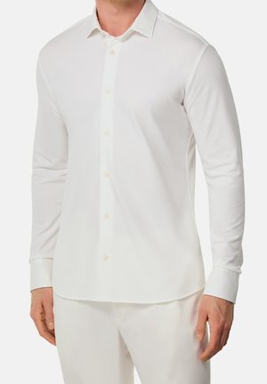 Homme portant une chemise blanche à manches longues boutonnée et un pantalon blanc, debout devant un fond clair uni.