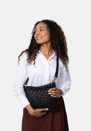 Borsa a tracolla in pelle nera intrecciata con chiusura a zip, tracolla regolabile e superficie testurizzata. Il modello tiene la borsa mentre indossa una camicia bianca.