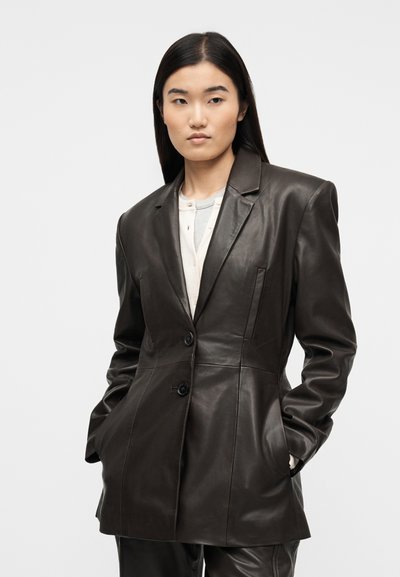Femme portant un blazer en cuir marron foncé à deux boutons, les mains dans les poches, associé à des hauts superposés de couleur claire en dessous.