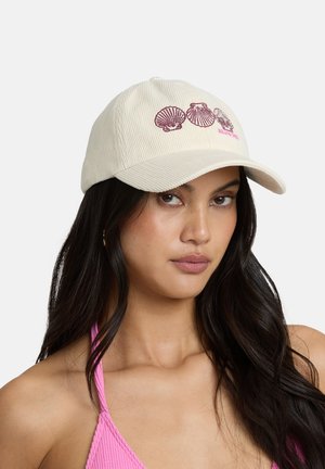 Woman with long dark hair wearing a cream corduroy cap embroidered with three seashells and a pink textured halter top.