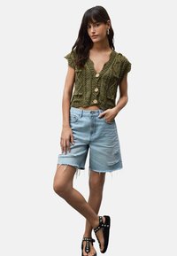 Vihreä neule crop-top, jossa on napit, yhdistettynä vaaleansinisiin farkkushortseihin, joissa on risat helmat ja kulutustekstuuria, lisättynä mustilla sandaaleilla.