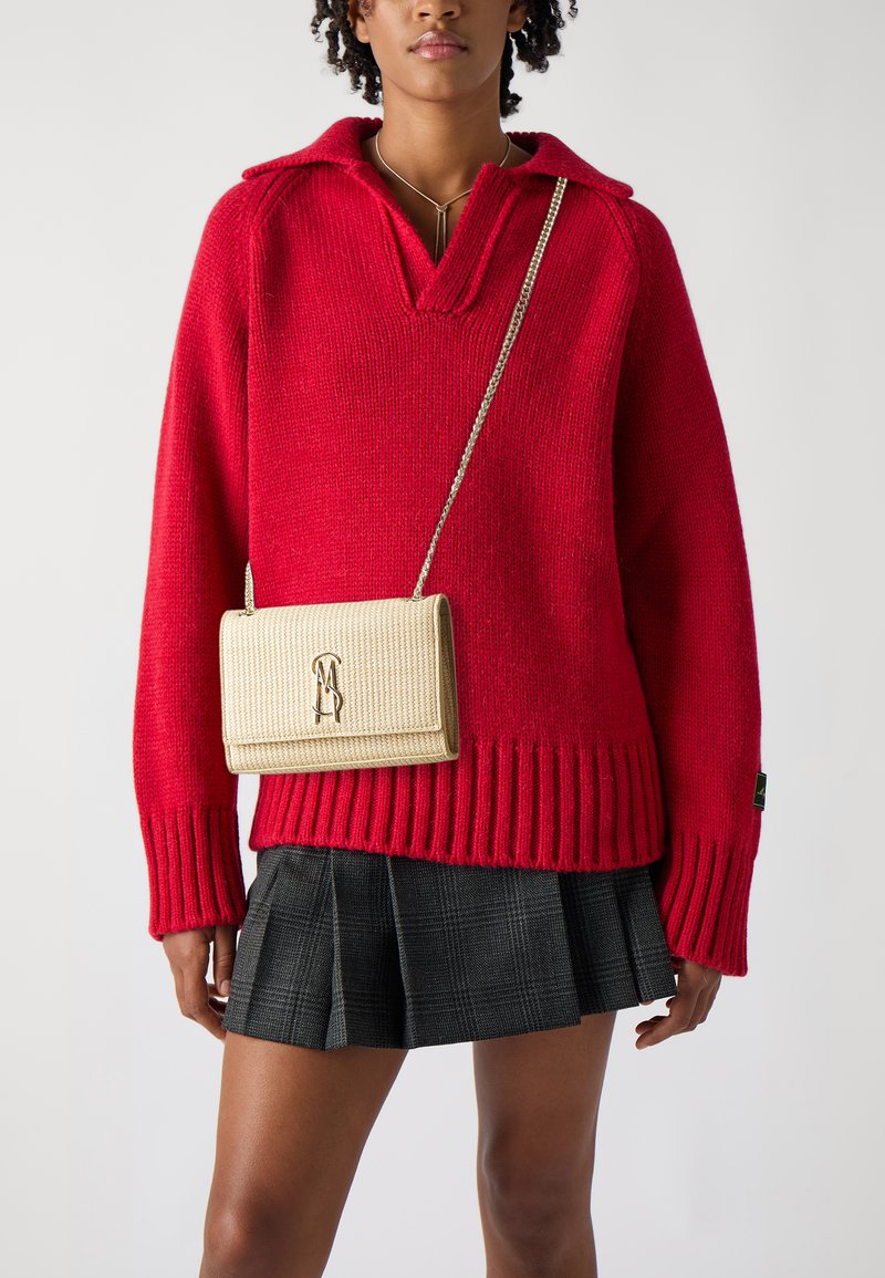 Červený pletený svetr s límcem a žebrovaným lemem, nošený přes černou kostkovanou plisovanou sukni. Malá béžová crossbody taška s detailním logem visí na řetízku.