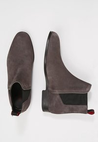 Bottines en daim gris à finition lisse, avec panneaux latéraux élastiques et un onglet accent rouge. Elles présentent une semelle plate noire et une forme d'orteil arrondie.