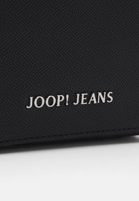 Čierna kožená peňaženka s embosovaným logom "JOOP! JEANS" v striebornej farbe. Má hladký povrch a šité okraje.