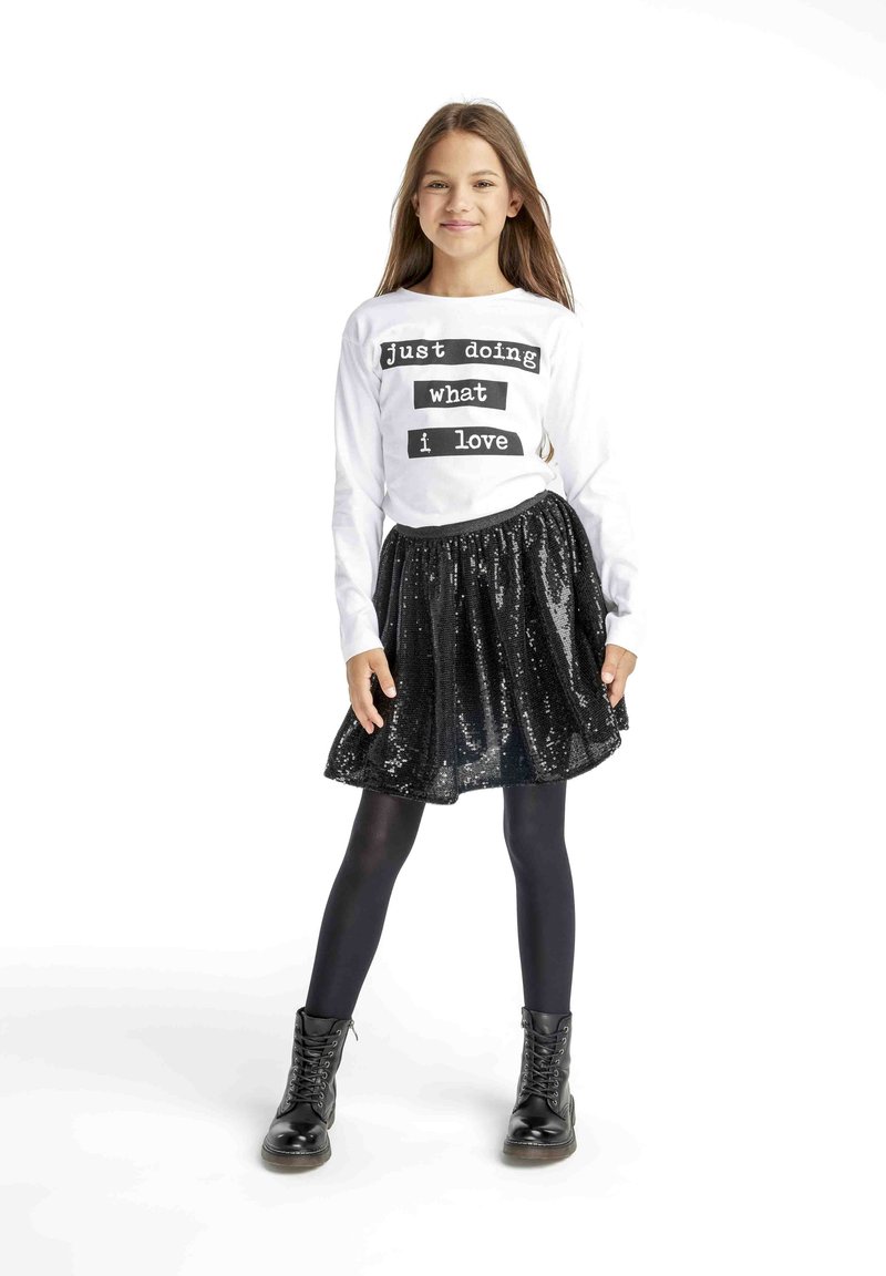 Witte shirt met lange mouwen en zwarte tekst, gecombineerd met een zwarte paillettenrok. Zwarte leggings en veterschoenen maken de outfit compleet.