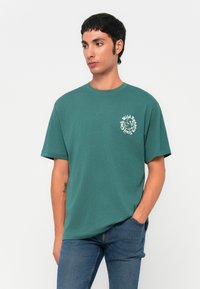 Camiseta de algodón de color verde azulado con mangas cortas y cuello redondo, que presenta una impresión gráfica blanca en el lado izquierdo del pecho. Combinada con jeans azules.