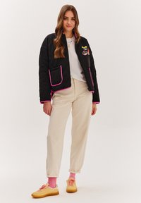 Čierna prešívaná bunda s ružovými akcentmi, prednými vreckami a farebnými výšivkami. Béžové nohavice a žlté tenisky s ružovými šnúrkami.