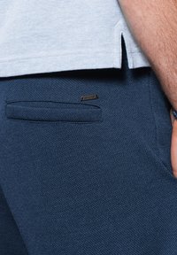 Modré kalhoty z texturované tkaniny s kapsou na zadní straně a malou logovou nášivkou. Světle modrý prodloužený lem košile překrývající pas.