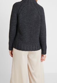 Tmavě šedý pletený svetr s vysokým límcem, žebrovanými manžetami a texturovaným vzorem. Kombinován se světlými béžovými kalhotami s širokými nohavicemi.