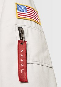Gray fabric with an American flag patch, metal zipper, and red fabric tag displaying "5.4.3.2.1." attached to a side pocket.