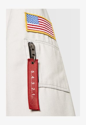 Siva tkanina s našitkom ameriške zastave, kovinskim zadrgo in rdečim tekstilnim oznako z napisom "5.4.3.2.1." pritrjeno na stranski žep.