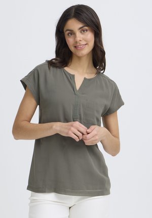 Jonge vrouw met donker haar, gekleed in een olijfgroene blouse met korte mouwen en witte broek, staand en glimlachend tegen een effen achtergrond.