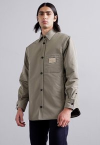 Jachetă cu buton în verde măslin, confecționată dintr-un material neted, având un singur buzunar pe piept cu etichetă și butoane negre. Design și croi simple.