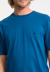 Blaues Baumwoll-T-Shirt mit rundem Halsausschnitt, kurzen Ärmeln und einem kleinen braunen Logo auf der linken Brustseite. Glatte Textur, lässige Passform.