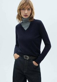Temno modra pletena majica z v-izrezom, kombinirana s sivo turtleneck majico. Model nosi temno jeans hlače in črn pas s srebrno sponko.