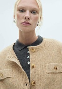 Beige stickad cardigan med ribbad struktur, med rund hals, träknappar och två bröstfickor över en mörk polotröja.