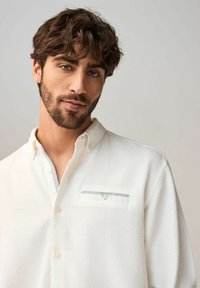 Chemise blanche à manches longues avec une finition texturée, dotée d'une petite poche poitrine avec des accents bleu clair et une fermeture à bouton.