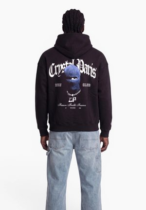 Man with braided hair wears black hoodie with blue ski mask graphic, "Crystal Paris Run Club" text, and light blue jeans, viewed from the back.