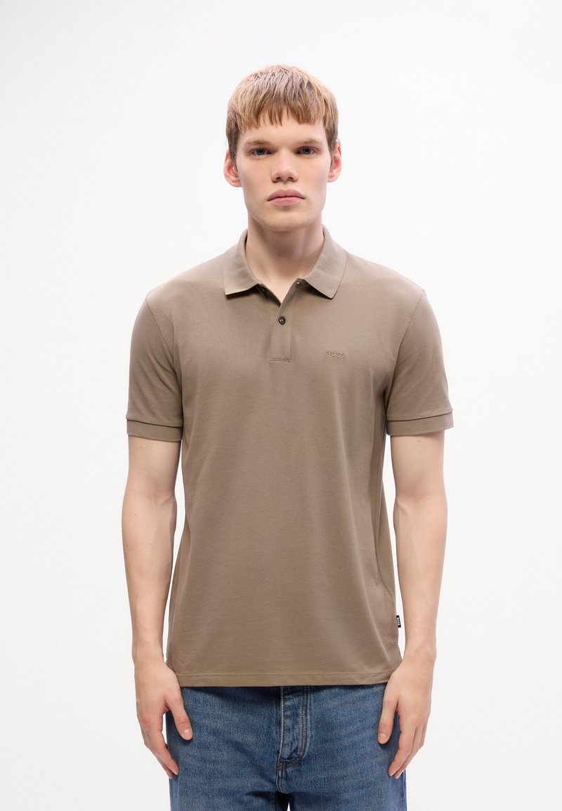 Jeune homme portant un polo taupe à manches courtes avec un petit logo "BOSS" et un jean bleu, se tenant devant un fond blanc uni.