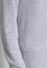 Pull gris clair à texture douce, avec ourlet et poignets côtelés, présentant des coutures discrètes le long des bras et du corps pour un look épuré.