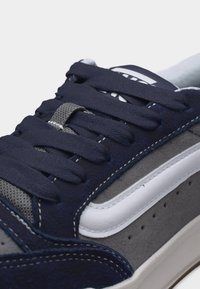 Κοντινό πλάνο γκρι και ναυτικού μπλε αθλητικού παπουτσιού με λευκή πλαϊνή ρίγα, χοντρές ναυτικές κορδόνια και λεπτομερείς ραφές σε σουέντ και δίχτυα υλικά.