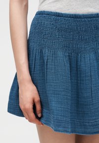 Μπλε φούστα με υφή και μαζεμένη ελαστική μέση, φορεμένη από άτομο με ανοιχτό γκρι τοπ, που δείχνει μέρος του χεριού και του μηρού.